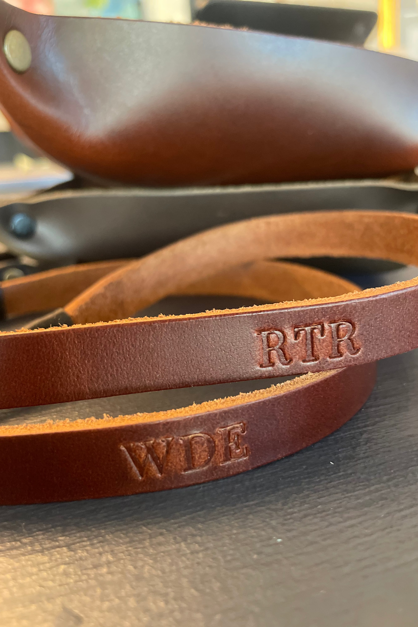 A brown leather sunglass strap with the monogram 'RTR' embossed on it, displayed on a table with a partial view of a smartphone case in the background.