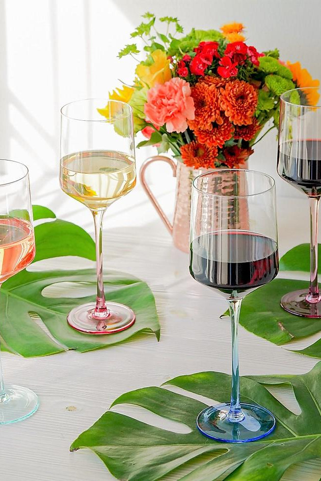 Four wine glasses with different types of wine on a table with floral decorations and large green leaves.